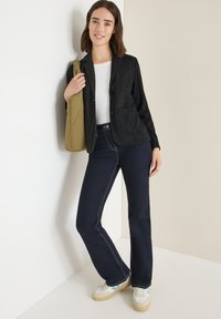 Sort tekstureret blazer over en hvid top, parret med mørkeblå flagrende jeans. Accessories inkluderer en blød oliven grøn taske og hvide sneakers.