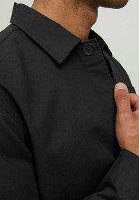 Sort button-up jakke med klassisk krave, glat stoftekstur og en lukning med knap på siden. Nærbillede af kraven og frontdetaljen.