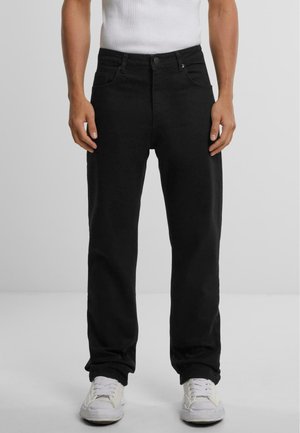 Mężczyzna ubrany w czarne jeansy o prostych nogawkach, białe trampki i białą bezrękawnik, stojący na tle jednolitego, jasnego tła.