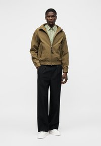 Homme portant une veste zippée vert olive par-dessus une chemise vert clair, un pantalon noir à jambes larges et des baskets blanches, debout devant un fond blanc.
