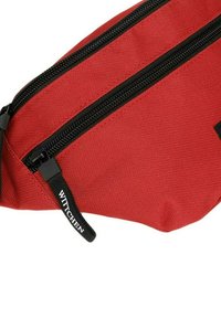 Rode stoffen heuptas met twee zwarte ritsen, gestructureerd oppervlak en een zwarte merk pull tab. Het ontwerp is compact en schuin.