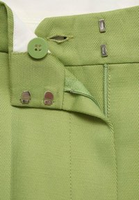 Close-up of green pants waistband showing button, zipper, and metal hook-and-eye fasteners on textured fabric.