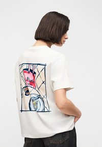 Γυναίκα με κοντά σκούρα μαλλιά, φορώντας λευκό μπλουζάκι με πολύχρωμο γραφικό του Παρισιού και σετ τραπεζιού στην πλάτη, συνδυασμένο με σκούρο τζιν.