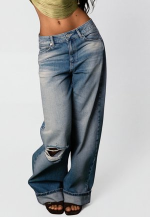 Osoba z widocznym brzuchem, ubrana w luźne, wyblakłe niebieskie dżinsy z podartym kolanem i szerokimi mankietami, do tego ciemne sandały z odkrytymi palcami na jasnym tle.