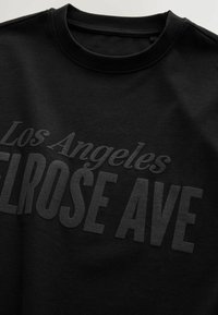 Crna pamučna majica s podignutim sivim natpisima "Los Angeles" i "Melrose Ave" različitim fontovima. Okrugli izrez i mekana tekstura.