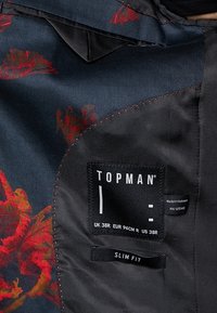 Tessuto nero con motivo floreale rosso, texture liscia. Presenta un'etichetta con "TOPMAN," dettagli sulla taglia e un'etichetta "SLIM FIT."