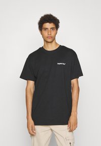 Sort bomulds t-shirt med korte ærmer, der har hvid tekst "glemte ansigtstræk" foran. Modellen bærer lysebeige bukser. Simpel rund halsudskæring.