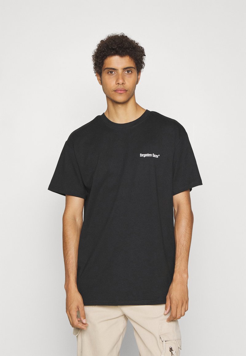 Sort bomulds t-shirt med korte ærmer, der har hvid tekst "glemte ansigtstræk" foran. Modellen bærer lysebeige bukser. Simpel rund halsudskæring.