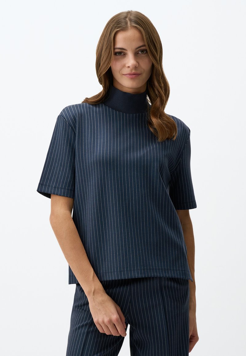 Granatowa bluzka z krótkim rękawem i wysokim kołnierzem, z pionowymi białymi prążkami. Miękka, gładka tkanina o luźnym kroju.