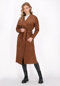 Brun ruskind lang frakke med bred krave, belagt talje og lige ærmer. Bæres over en lys top og kombineres med sorte støvler og jeans.