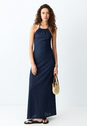 Donna con capelli ricci che indossa un vestito lungo a collo halter blu navy, tiene una piccola borsa di paglia e indossa sandali blu navy.