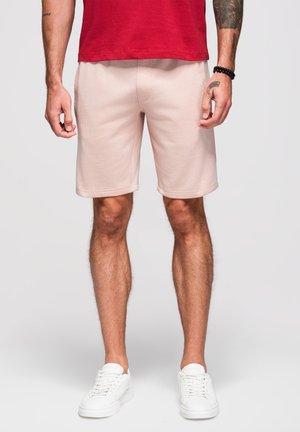 Homme portant un short rose clair, un t-shirt rouge, des baskets blanches et un bracelet noir en perles, debout devant un fond uni.