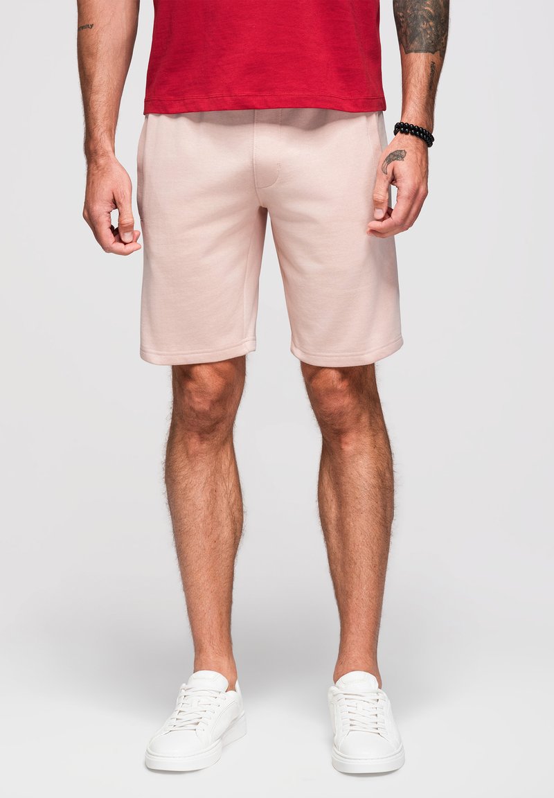 Man draagt lichtroze korte broek, rood t-shirt, witte sneakers en een zwarte kralenarmband, staand tegen een effen achtergrond.