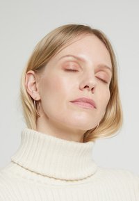 Modèle portant un pull côtelé couleur crème avec une texture douce et un design épuré. De simples boucles d'oreilles créoles complètent le look.