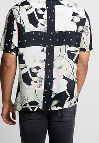 Krátký rukáv košile s barevně blokovaným designem, který obsahuje květinové vzory, hvězdy a směs černé, zelené a pastelových barev.