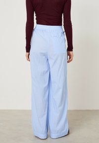 Šviesiai mėlyni dryžuoti platėjančių klešnių kelnės su elastiniu liemeniu ir atvira kišene nugaroje. Lygus audinys, laisvo silueto, rodomos iš nugaros.