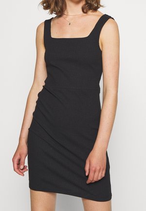 Kobieta w czarnej, prążkowanej, dzianinowej sukience bez rękawów o kwadratowym dekolcie oraz delikatnym naszyjniku z małym wisiorkiem.