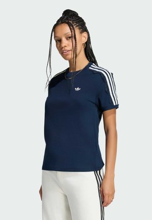 Kvinde med flettet hår bærer en mørkeblå Adidas t-shirt med hvide striber og hvide bukser med sorte sidelister mod en lys baggrund.