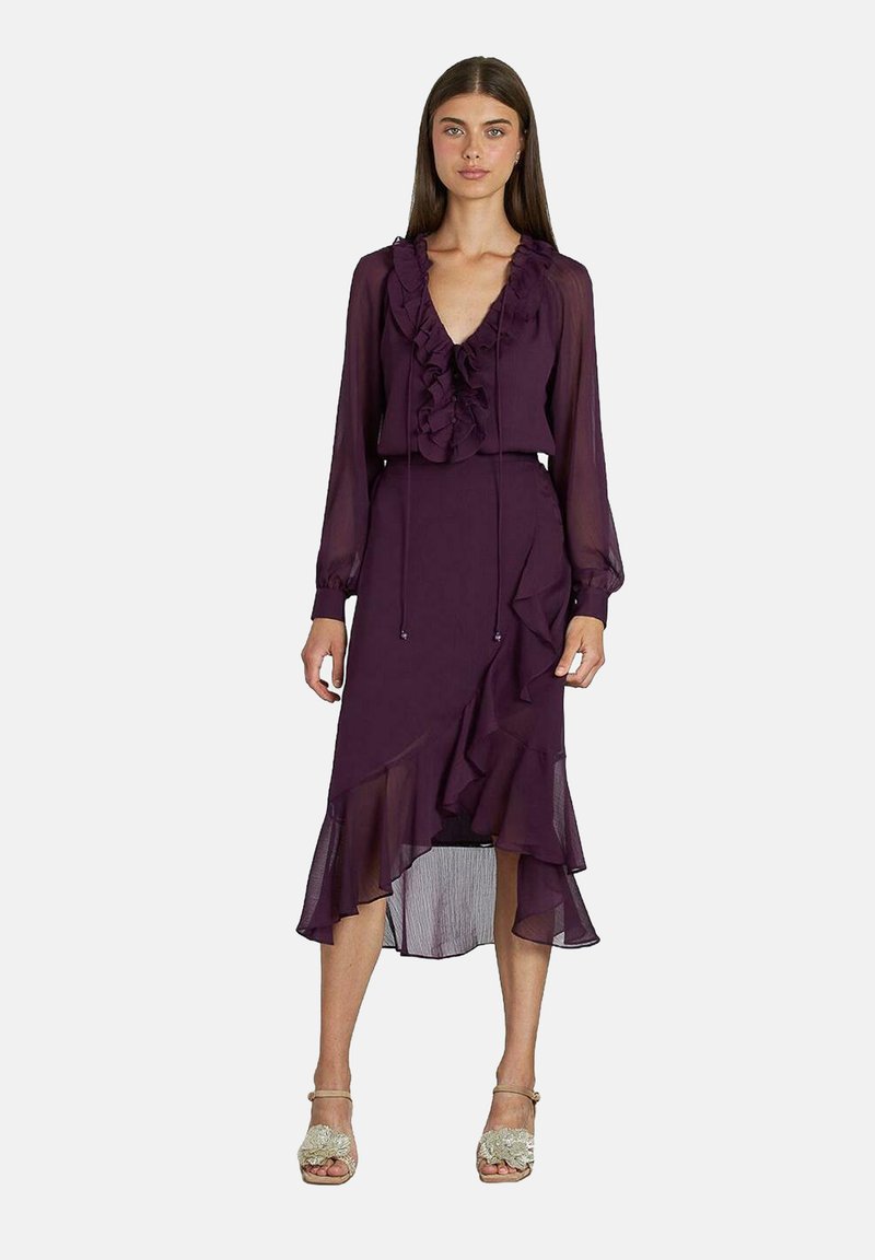 Robe violette avec un col froncé et des manches longues, présentant un ourlet à volants. Fabriquée en tissu léger avec une texture fluide.
