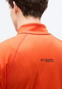 Κοντομάνικο πολο σε πορτοκαλί χρώμα Columbia Titanium, με υφασμάτινη υφή και διακριτικό σχέδιο, με μαύρο λογότυπο στην πλάτη.