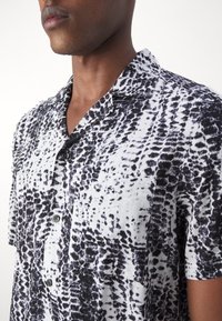Homme portant une chemise à manches courtes à boutons avec col, noire et blanche à motif abstrait, légèrement tourné vers la gauche sur un fond uni.
