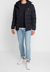 Giacca a vento nera con cappuccio, con foderatura bordeaux, tasche con zip e un accento di logo. Indossata sopra un felpa nera e jeans azzurri chiari.