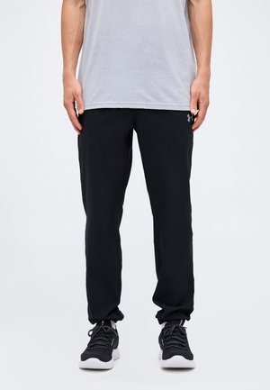 Άτομο στέκεται φορώντας μαύρο παντελόνι Under Armour, γκρι μπλουζάκι και μαύρα αθλητικά παπούτσια με λευκές σόλες σε ένα απλό φωτεινό φόντο.