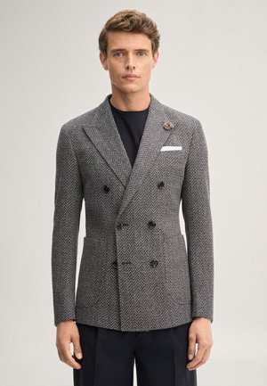Man draagt een grijze visgraat dubbelgeknoopt colbert met een zwart overhemd en donkere broek tegen een effen achtergrond.