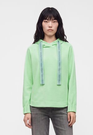 Ung kvinde med langt sort hår iført en lys grøn hættetrøje med brede snore og grå jeans, står mod en ensfarvet hvid baggrund.
