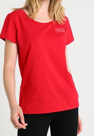 Kvinde iført en ensfarvet rød T-shirt med korte ærmer med en lille logo på øverste venstre bryst og sorte bukser.