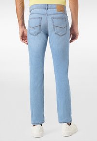 Man som bär ljusblå raka jeans och vita sneakers, står med ryggen mot kameran mot en vit bakgrund.