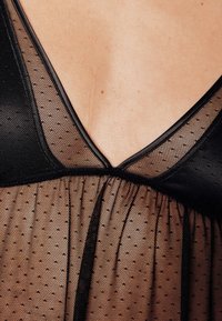 Tissu noir transparent à petits pois, avec un décolleté en V profond et des fronces élastiques à la taille. Les bords sont finis par un liseré noir.