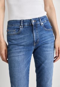 Ljusblå denimjeans med midja i normalhöjd, synliga stygn, framfickor och knappstängning vid midjan.