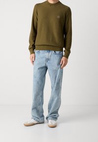 Pull à côtes vert olive avec un col rond, associé à un jean bleu clair à coupe ample et des baskets blanches avec des semelles brunes.