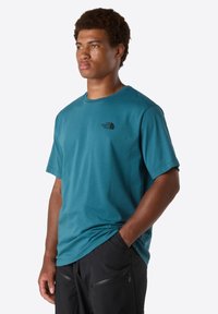 Joven con cabello rizado que lleva una camiseta teal de The North Face y pantalones negros con cremalleras, mirando hacia la izquierda con una mano en el bolsillo.