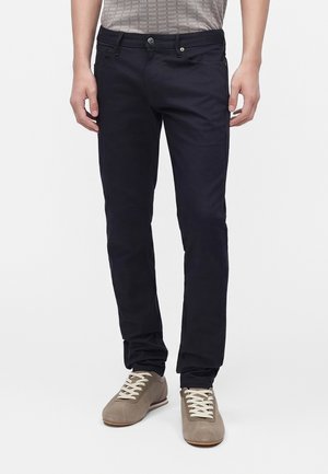 Uomo con pantaloni neri slim fit e sneaker casual marrone chiaro, in piedi con le braccia rilassate lungo i fianchi su uno sfondo semplice.
