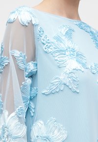 Tissu bleu clair transparent avec une grande broderie florale en relief dans un bleu assorti couvrant l'épaule et le bras.