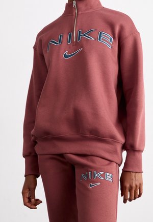 Osoba nosząca pasujący do siebie różany brudny dres Nike z logo i znakiem na klatce piersiowej i udzie, stojąca z rozluźnionymi ramionami.