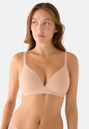 Joven mujer con un sujetador nude, ligeramente acolchado y sin aros, con tirantes finos, mirando ligeramente hacia su izquierda sobre un fondo liso.