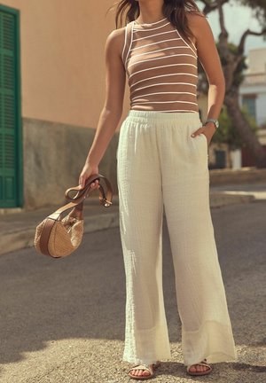 Femme portant un haut sans manches marron à rayures et un pantalon blanc ample, tenant un sac à main tissé avec des lanières en cuir, debout en plein air dans une rue ensoleillée.