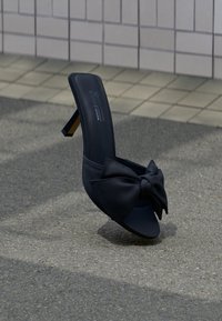 Πέδιλο mule μαύρο με ψηλό τακούνι και μεγάλο φιόγκο στο μπροστινό μέρος, τοποθετημένο σε υφή πεζοδρομίου κοντά σε φόντο τοίχου με πλακάκια.