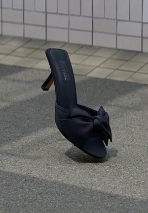 Sandale à talon haut noire en cuir suédé avec grand nœud sur l'avant-pied, posée sur un pavé texturé près d'un mur carrelé en arrière-plan.