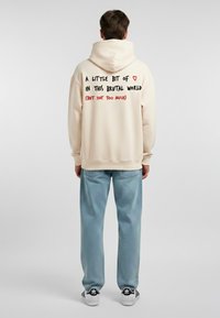 Felpa con cappuccio beige dalla vestibilità rilassata. Grafica testuale sulla schiena in nero e rosso. Pantaloni di jeans azzurri chiari e sneaker in bianco e nero.