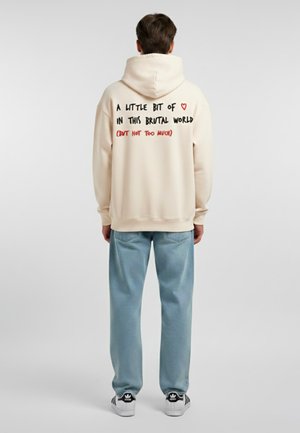 Felpa con cappuccio beige dalla vestibilità rilassata. Grafica testuale sulla schiena in nero e rosso. Pantaloni di jeans azzurri chiari e sneaker in bianco e nero.