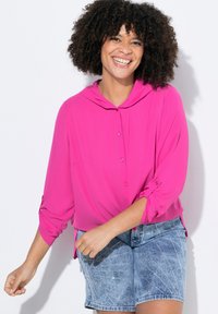 Rosa Bluse mit Knopfleiste und Kapuze, lockere Passform und hochgekrempelten Ärmeln, kombiniert mit hellblauen Jeansshorts mit dekorativen Nähten.