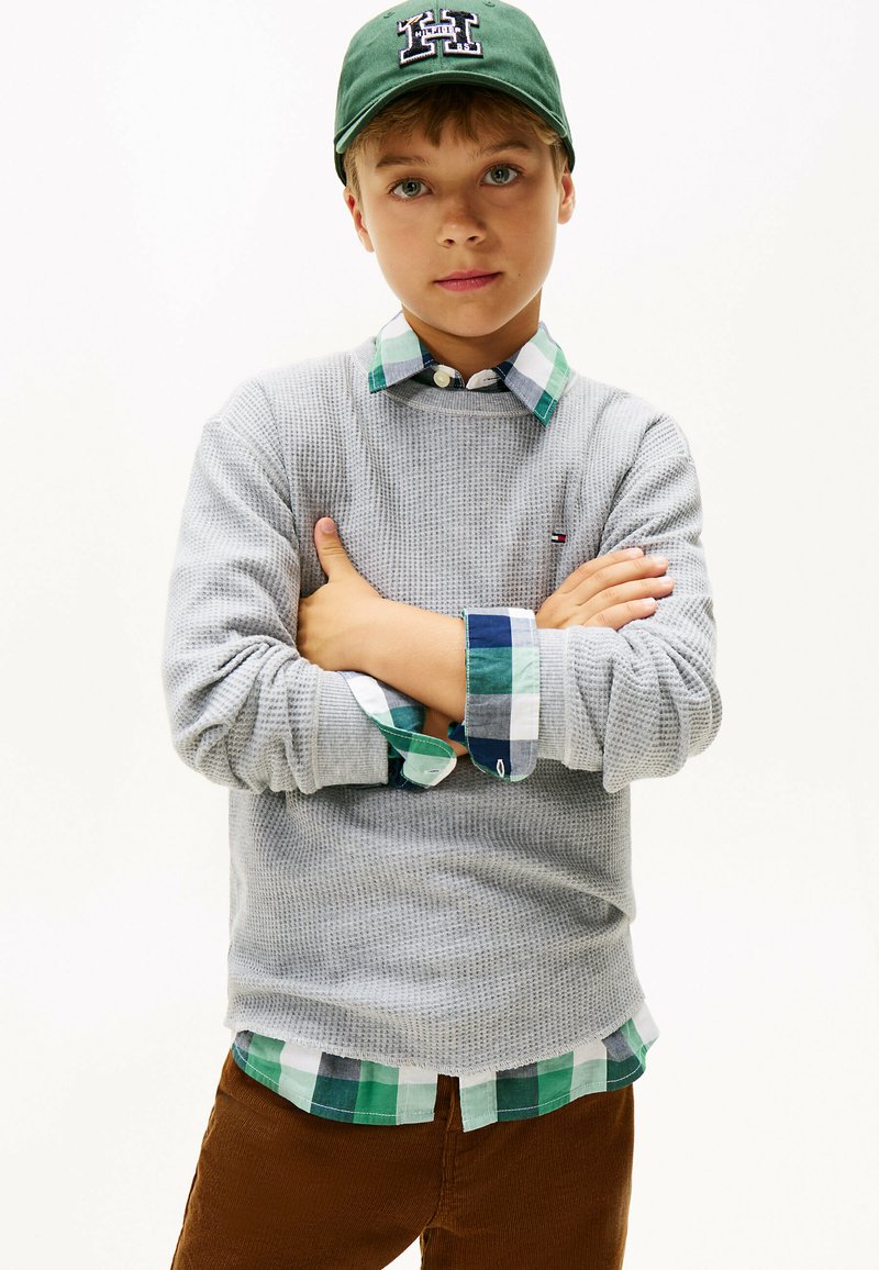 Maglione grigio testurizzato indossato sopra una camicia a quadri verde e blu. Il soggetto indossa un berretto verde con un logo letterato. Pantaloni in velluto a coste marroni.