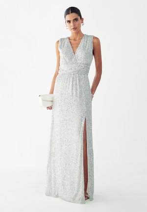 Kvinde iført ærmeløs sølv palietkjole med lårslids, der holder en hvid clutch, stående foran en ensfarvet hvid baggrund.