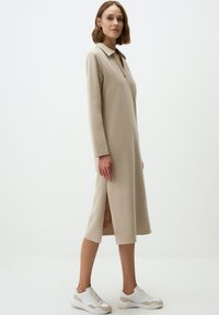Robe midi beige avec un col, des manches longues et des fentes sur les côtés. Confectionnée en tissu texturé. Portée avec des baskets blanches et beiges.