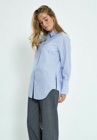 Camicia button-up blu chiaro con strisce verticali bianche e arancioni, dotata di maniche lunghe e orlo arrotondato, abbinata a pantaloni grigi.