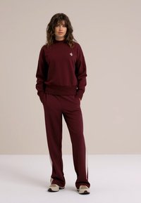 Burgundi pulóver és hozzá illő nadrág, puha anyagból készült. A pulóver bordás mandzsettákkal és egy kis logóval rendelkezik; a nadrág kontrasztos oldalsávokkal díszített.